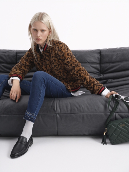 Model sitting on grey sofa wearing animal-print jacket, embellished jeans, white socks and black loafers; green handbag and sunglasses beside her.
