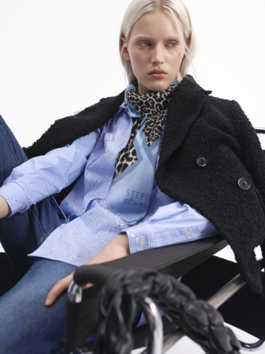Model seated in chair wearing a STEFFEN SCHRAUT outfit with light blue shirt, jeans, black jacket and printed scarf.