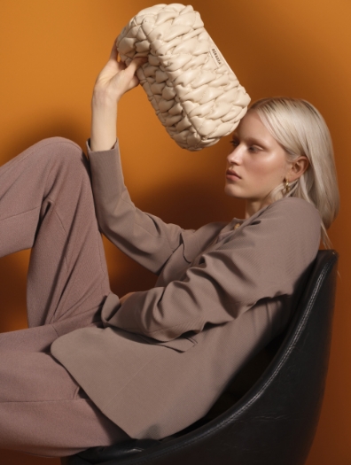 Model wearing a taupe-colored STEFFEN SCHRAUT suit, holding a woven beige handbag against an orange background.
