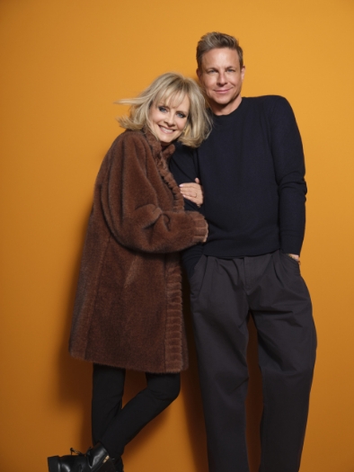 Twiggy leans on Steffen Schraut, both smiling in front of an orange background – a portrait of fashion icons.